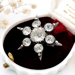 May include: A vintage starburst brooch featuring clear, faceted gemstones. The brooch is set in silver-toned metal and presented in a white jewellery box with a red velvet interior. The brooch has a central large stone surrounded by smaller stones.