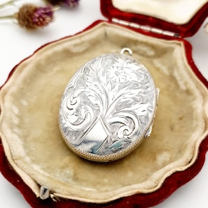 Peut inclure: Médaillon ovale en argent avec un motif floral gravé, présenté dans un écrin en velours rouge. Le médaillon est ouvert, révélant un intérieur clair. Le motif floral comprend des feuilles et une fleur centrale.