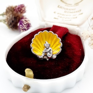 May include: A vintage silver-tone ring featuring a seated elf figure within a yellow enamel shell design. The ring is presented in a white jewelry box with a red velvet interior, showcasing the ring's intricate details and craftsmanship.