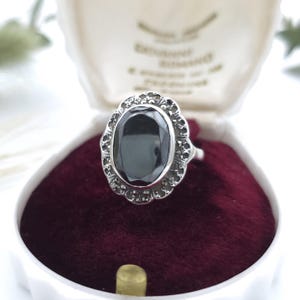 May include: A silver ring featuring a large, oval-shaped, faceted black stone set in a scalloped frame adorned with small, dark, metallic accents. The ring is displayed inside a white, clam-shaped jewellery box with a deep red velvet interior lining.