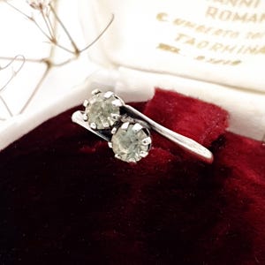 May include: A silver ring with two clear stones set in a prong setting. The ring is on a red velvet background.