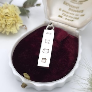 May include: A rectangular silver pendant featuring four distinct hallmarks stamped vertically along its front surface. The pendant is displayed inside a white jewellery box lined with deep burgundy velvet, which includes printed text on the lid.