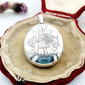 May include: Oval silver locket with an engraved monogram design. The locket is displayed open in a red velvet lined box. The locket has a loop at the top for a chain. The background is a light beige color.