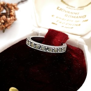 Könnte beinhalten: Ein silberner Ring mit einem Band aus kleinen, runden, hellgrünen Edelsteinen. Der Ring befindet sich in einer samtgefütterten Schatulle mit dem Text "GIOVANNI ROMANO" auf der Innenseite.