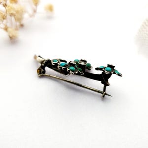 May include: A silver-toned brooch with a floral design. The brooch features small, round turquoise stones set in a dark metal frame, with green accents. The pin is a delicate piece of jewelry.