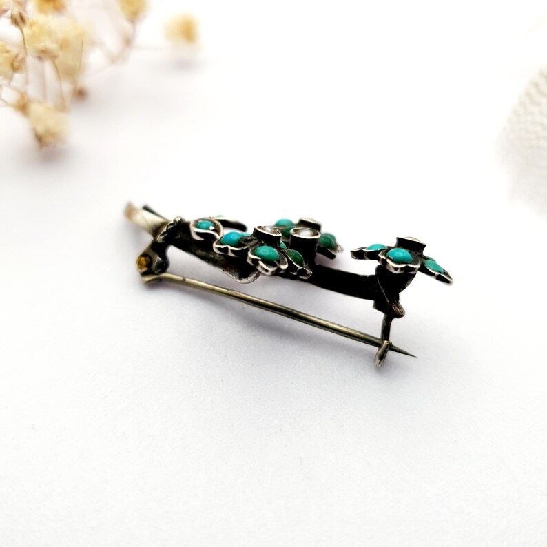 May include: A silver-toned brooch with a floral design. The brooch features small, round turquoise stones set in a dark metal frame, with green accents. The pin is a delicate piece of jewelry.