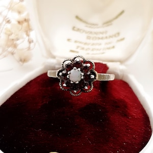 May include: A vintage ring featuring a flower design, with a central opal and surrounding dark red garnet stones. The ring is set in silver-toned metal. The ring is displayed on a red velvet surface.
