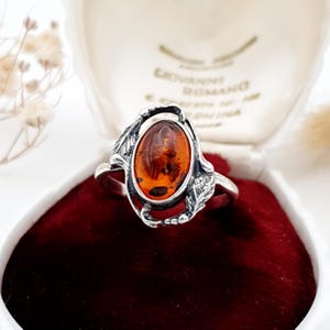 May include: A silver ring featuring an oval-shaped, translucent amber gemstone set in a decorative metal frame with leaf-like details. The band is polished silver, and the setting surrounds the warm, orange-brown stone with intricate metalwork.