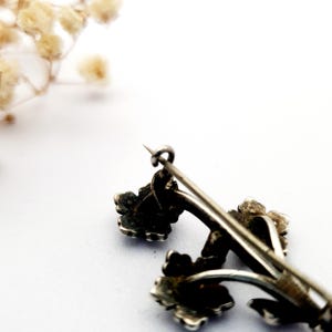 May include: A vintage silver-toned brooch featuring a floral design. The brooch has three flower clusters with detailed petals and stems. The pin is slightly tarnished, giving it an antique appearance. The background is white with a few dried flowers.