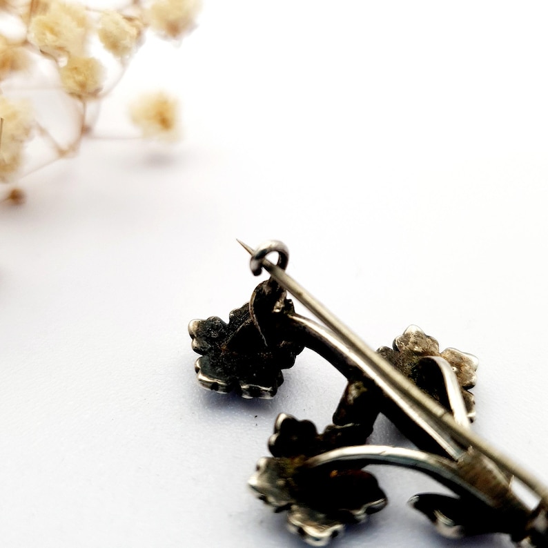May include: A vintage silver-toned brooch featuring a floral design. The brooch has three flower clusters with detailed petals and stems. The pin is slightly tarnished, giving it an antique appearance. The background is white with a few dried flowers.
