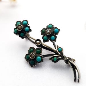 May include: A silver-toned floral brooch featuring three flower clusters with turquoise and green gemstone petals and a central pearl. The brooch has a stem and leaf design, with additional turquoise accents. The brooch is set against a white background.