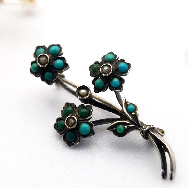 May include: A silver-toned floral brooch featuring three flower clusters with turquoise and green gemstone petals and a central pearl. The brooch has a stem and leaf design, with additional turquoise accents. The brooch is set against a white background.