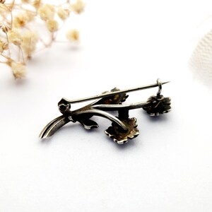May include: A silver-toned brooch in the shape of a floral sprig. The brooch features delicate leaves and three small, detailed flower blossoms. The pin is designed with a classic, elegant style, perfect for adding a touch of vintage charm to any outfit.