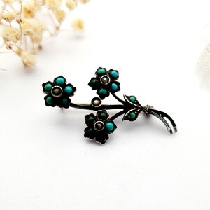 May include: A silver-toned floral brooch featuring three flower clusters with turquoise-colored stones and a central pearl. The brooch has a delicate, detailed design, with leaves and stems. The brooch is set against a white background.