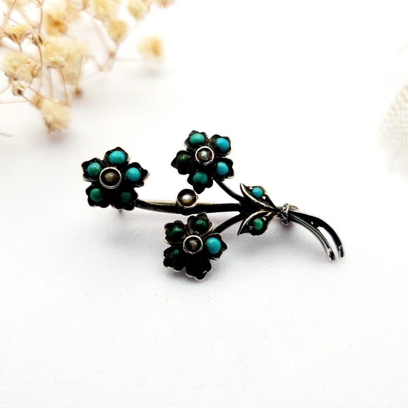 May include: A silver-toned floral brooch featuring three flower clusters with turquoise-colored stones and a central pearl. The brooch has a delicate, detailed design, with leaves and stems. The brooch is set against a white background.