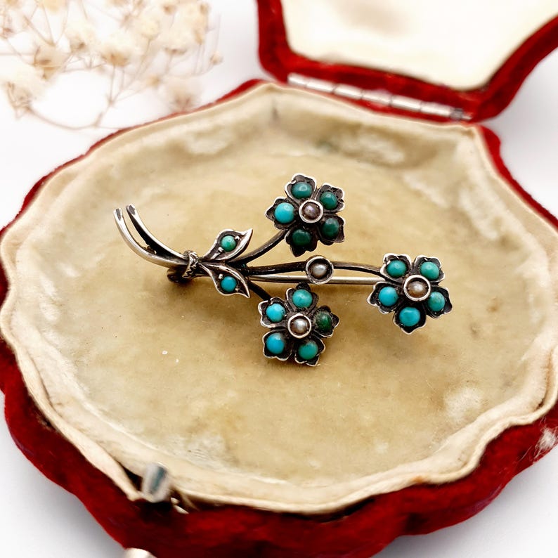May include: A silver-toned floral brooch with turquoise and green stone accents. The brooch features three flower clusters and small leaves, set against a red velvet jewelry box.