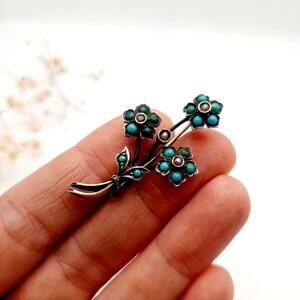 May include: A silver-toned floral brooch featuring three flower clusters with turquoise and green gemstone accents. The brooch has a delicate, detailed design with small leaves and pearl-like embellishments, creating an elegant, vintage aesthetic.
