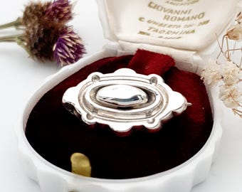 May include: A silver brooch with an oval center and a decorative border, presented in a white jewelry box with a red velvet interior. The box is open, revealing the brooch. Dried flowers are in the background.