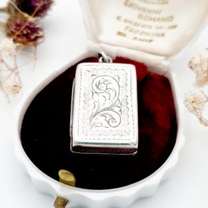 May include: A rectangular silver pendant with an engraved floral design, presented in a white box with a red velvet lining. The pendant has a small loop at the top. The box is open, displaying the pendant.