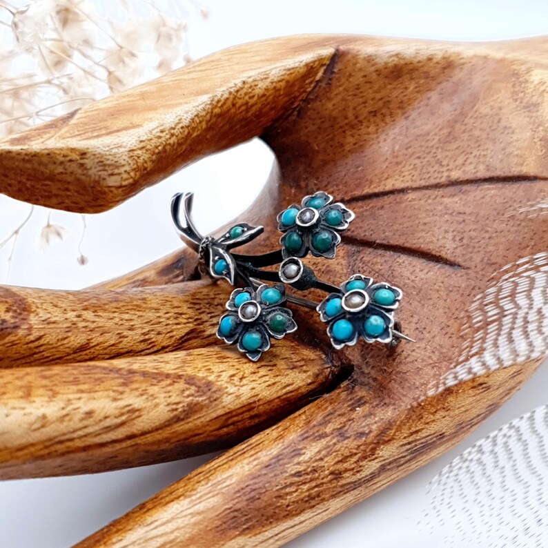 May include: A silver-toned floral brooch featuring three flower clusters with turquoise-colored stones and silver accents. The brooch has a delicate, detailed design, with a pin on the back for secure fastening. The brooch is a vintage-inspired accessory.