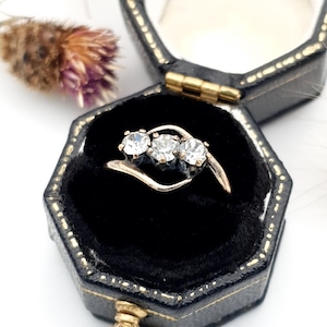 May include: A gold ring with three clear gemstones set in a curved design, displayed in a black velvet jewelry box. The box is hexagonal with gold trim. A dried flower with purple and brown hues is in the background.