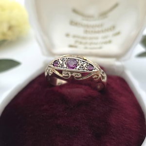 May include: A silver-toned ring featuring three round, deep pink stones set in a decorative metal band with intricate scrollwork details. The ring is positioned inside a white jewelry box with a dark red velvet interior lining.