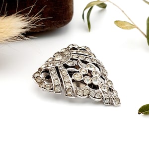 May include: A silver-coloured, vintage brooch with a leaf design. The brooch is adorned with many clear, sparkling rhinestones. The intricate details and arrangement of the stones create a dazzling effect, perfect for adding a touch of glamour.