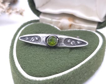 May include: A silver-toned metal brooch featuring an elongated, pointed shape with a central round, faceted lime-green stone. The metal surface displays engraved leaf motifs on both sides of the stone, set within a heart-shaped green velvet box.