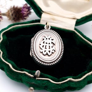 May include: An oval silver locket with a decorative design inside, set in a green velvet box. The locket has a beaded border and a small loop at the top for a chain. The box is open, revealing the locket against a dark background.
