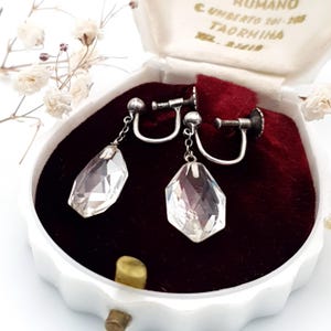 Op de afbeelding: Een paar vintage kristallen oorhangers, gepresenteerd in een witte sieradendoos met een rode fluwelen binnenkant. De oorbellen hebben een zilverkleurige metalen zetting en een facetgeslepen, druppelvormig kristal. Delicate witte bloemen op de achtergrond.
