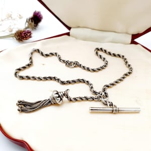 May include: A silver-toned necklace with a rope chain design and a tassel pendant. The necklace features a T-bar clasp and is displayed in a red-trimmed, cream-colored box. The tassel is made of multiple strands.