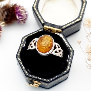 May include: Silver ring with an oval amber stone, presented in a black jewellery box. The ring has Celtic knot designs. The amber stone is a warm, golden-brown colour.