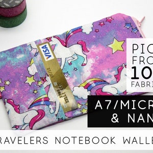 Könnte beinhalten: Ein Travelers Notebook Wallet mit einem bunten Einhorn- und Regenbogenmuster. Die Geldbörse hat einen rosa Reißverschluss und ein rosa Gummiband. Der Text auf dem Bild lautet "Pick from 100 Fabrics" und "A7/Micro & Nano". Eine Kreditkarte ist sichtbar.