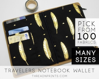 May include: A black travelers notebook wallet with a gold feather pattern and a zipper closure. The wallet has multiple card slots and a pocket for cash. The text "PICK FROM 100 FABRICS" and "MANY SIZES" is printed on the right side of the image. The text "TRAVELERS NOTEBOOK WALLET" and "THREADNPRINTS.COM" is printed at the bottom of the image.
