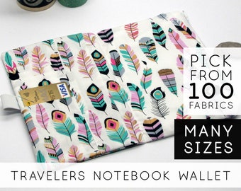 May include: A white fabric notebook wallet with a colorful feather pattern. The wallet has a pocket for a credit card. The text "PICK FROM 100 FABRICS" and "MANY SIZES" is visible in the image. The text "TRAVELERS NOTEBOOK WALLET" is visible at the bottom of the image.