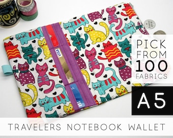May include: A fabric A5 travelers notebook wallet featuring a white background with a colorful pattern of cats in shades of yellow, teal, pink, and purple, accented by small black hearts. It includes a purple vertical card slot section.