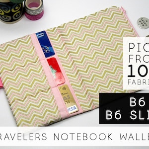 May include: A pink and gold chevron patterned fabric wallet with a zipper closure. The wallet has multiple card slots and a pocket for cash. The text "PICK FROM 100 FABRICS" and "B6 & B6 SLIM" is printed on the image. The text "TRAVELERS NOTEBOOK WALLET" is printed at the bottom of the image.