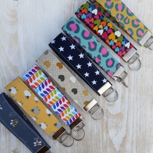 May include: A selection of fabric wrist keychains featuring diverse patterns such as bees, sheep, leaves, hearts, stars, and leopard print. Each keychain has a metal clasp and a key ring. The keychains are displayed diagonally on a light wooden surface.