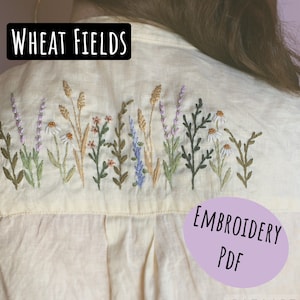 Op de afbeelding: Crèmekleurig overhemd met een geborduurd bloemmotief. De borduursels bevatten tarwe, lavendel, madeliefjes en andere wilde bloemen in groen, paars en bruin. Tekst op het shirt luidt "WHEAT FIELDS" en "EMBROIDERY PDF".