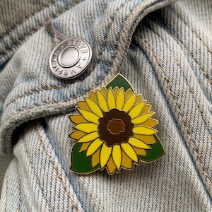 May include: A sunflower enamel pin with yellow petals, a brown centre, and green leaves. The pin is set against a light blue denim jacket, with a silver button visible in the background. The pin is a decorative accessory.