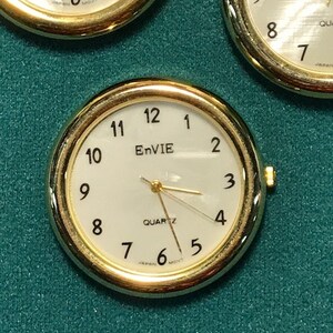 May include: Close-up of three gold-toned quartz watch faces with white dials. The watches have black numerals and hands, with the brand name "EnVIE" visible on one. The background is a textured teal surface.
