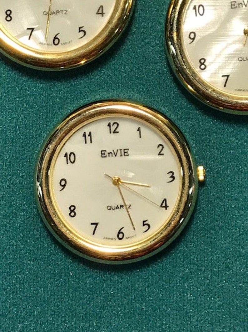 May include: Close-up of three gold-toned quartz watch faces with white dials. The watches have black numerals and hands, with the brand name "EnVIE" visible on one. The background is a textured teal surface.