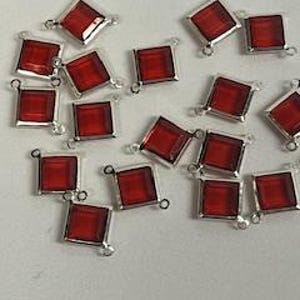 Op de afbeelding: Een groep van 18 rode vierkante glazen kralen met zilverkleurige metalen lussen aan elke kant. De kralen zijn willekeurig gerangschikt op een wit oppervlak.