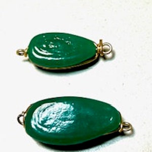 May include: Two green teardrop-shaped pendants with gold-colored wire loops. The pendants are smooth and glossy, with a slight variation in size. The background is a light, neutral color.