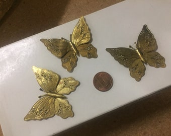 May include: Three gold-colored metal butterflies with intricate designs. The butterflies are about the size of a US penny.