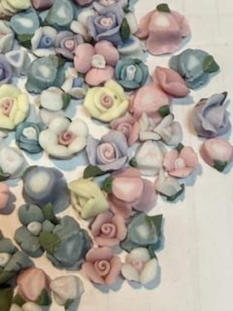 May include: Assortment of pastel colored porcelain flower embellishments. The flowers are in various shapes and sizes, including roses, daisies, and other floral designs.