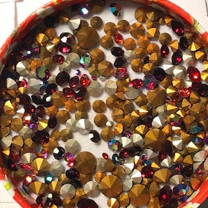 May include: A round container filled with a variety of small, colorful rhinestones. The rhinestones are in shades of gold, red, and iridescent.