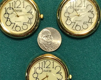 May include: Three gold-tone watch faces with white dials and black numbers. The dials read "EnVie Quartz".