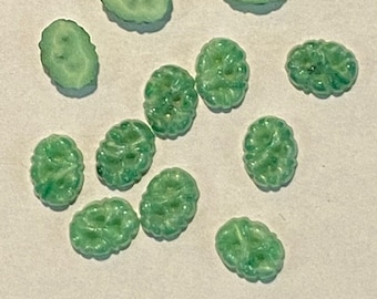 Puede incluir: Una colección de cuentas de jade verde en varias formas, incluyendo diseños ovalados y florales. Las cuentas están dispersas sobre una superficie clara, mostrando su textura pulida y color vibrante. Adecuado para la creación de joyas.