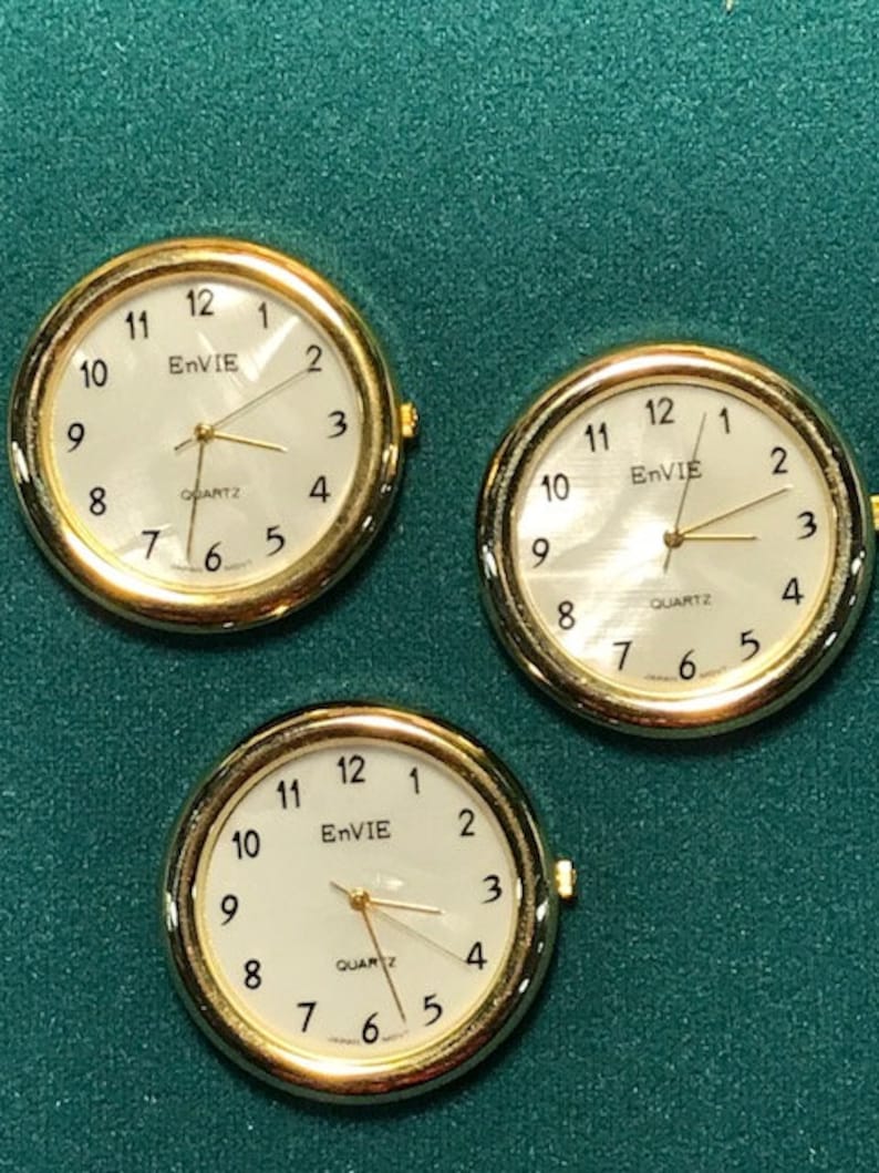 May include: Three gold-toned quartz watches with white faces and black numerals. The watch faces display the brand name "EnVIE" and the word "QUARTZ". The hands are gold-toned, and the watches are set against a green background.